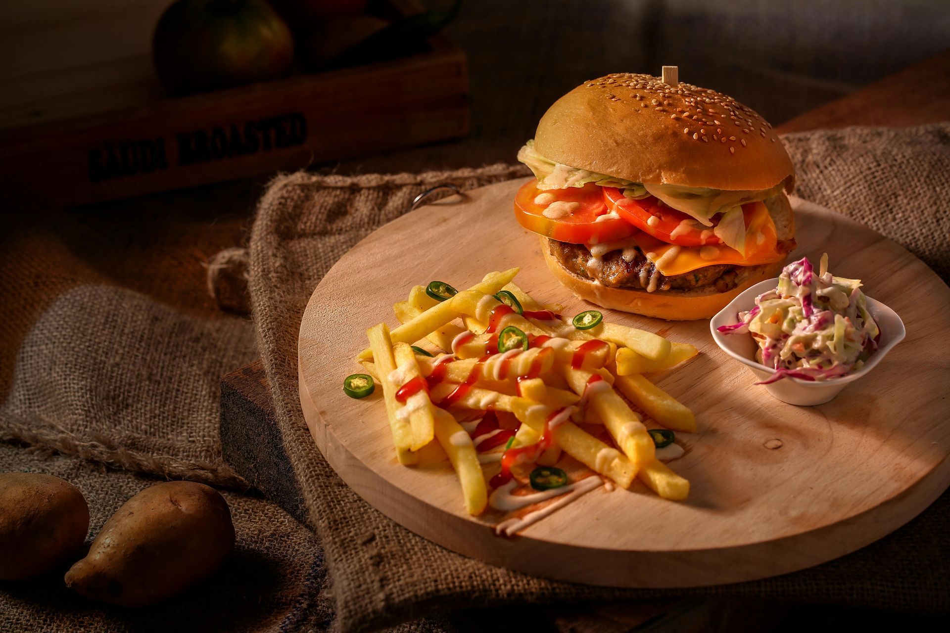 A sesame seed bun burger with fries topped with red sauce and green peppers, served with coleslaw on a wooden board.