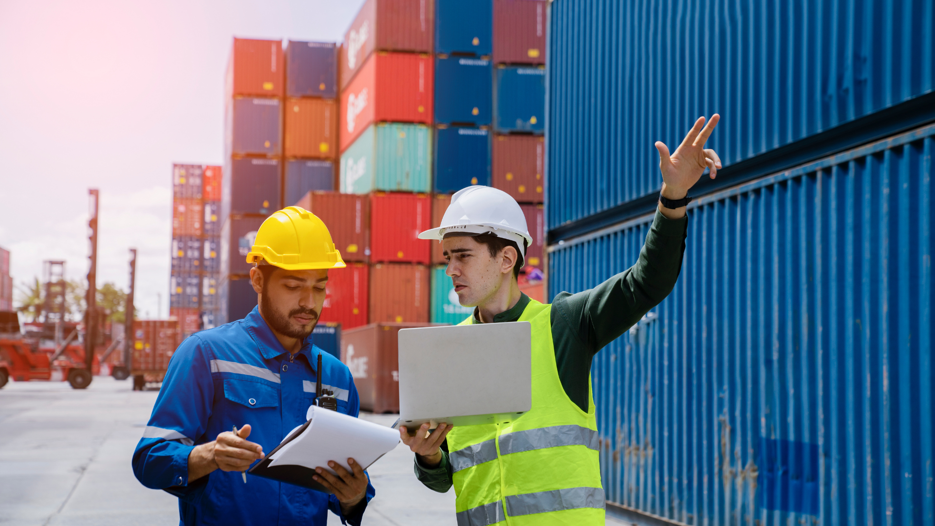 Two men, wearing hard hats and safety vests, in a shipping yard reviewing paperwork and using a laptop next to shipping containers.