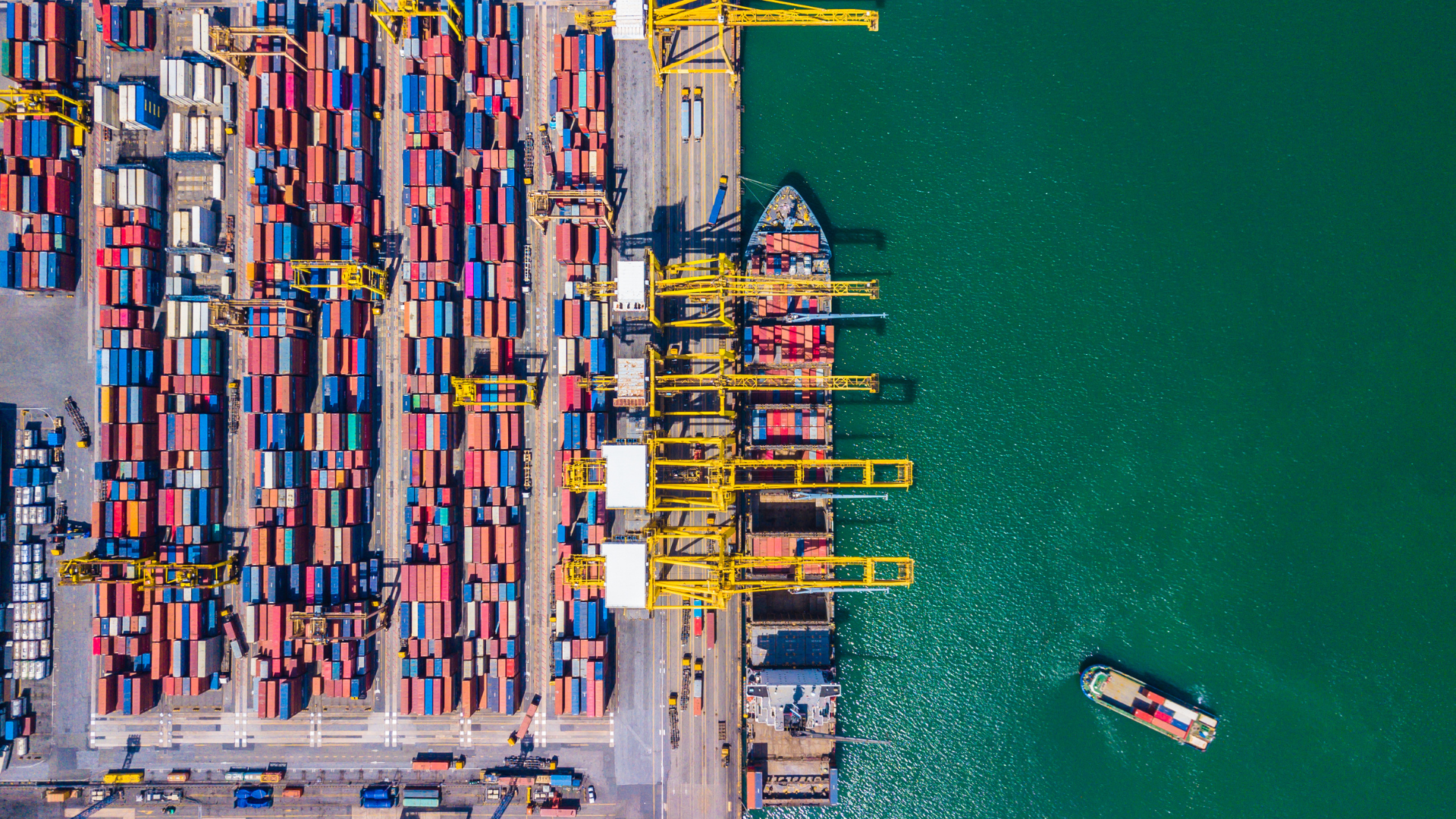 Aerial view of a busy shipping port with container stacks, cranes, and ships; turquoise water.