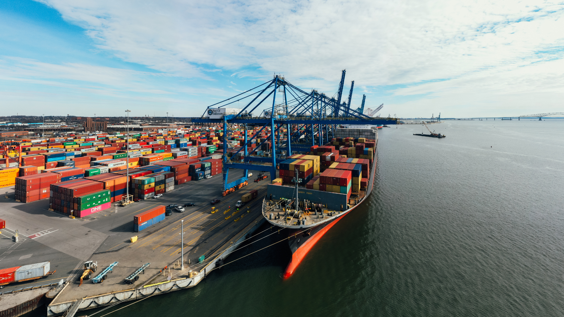 Cargo ship docked at a port, being unloaded by large cranes, surrounded by shipping containers.
