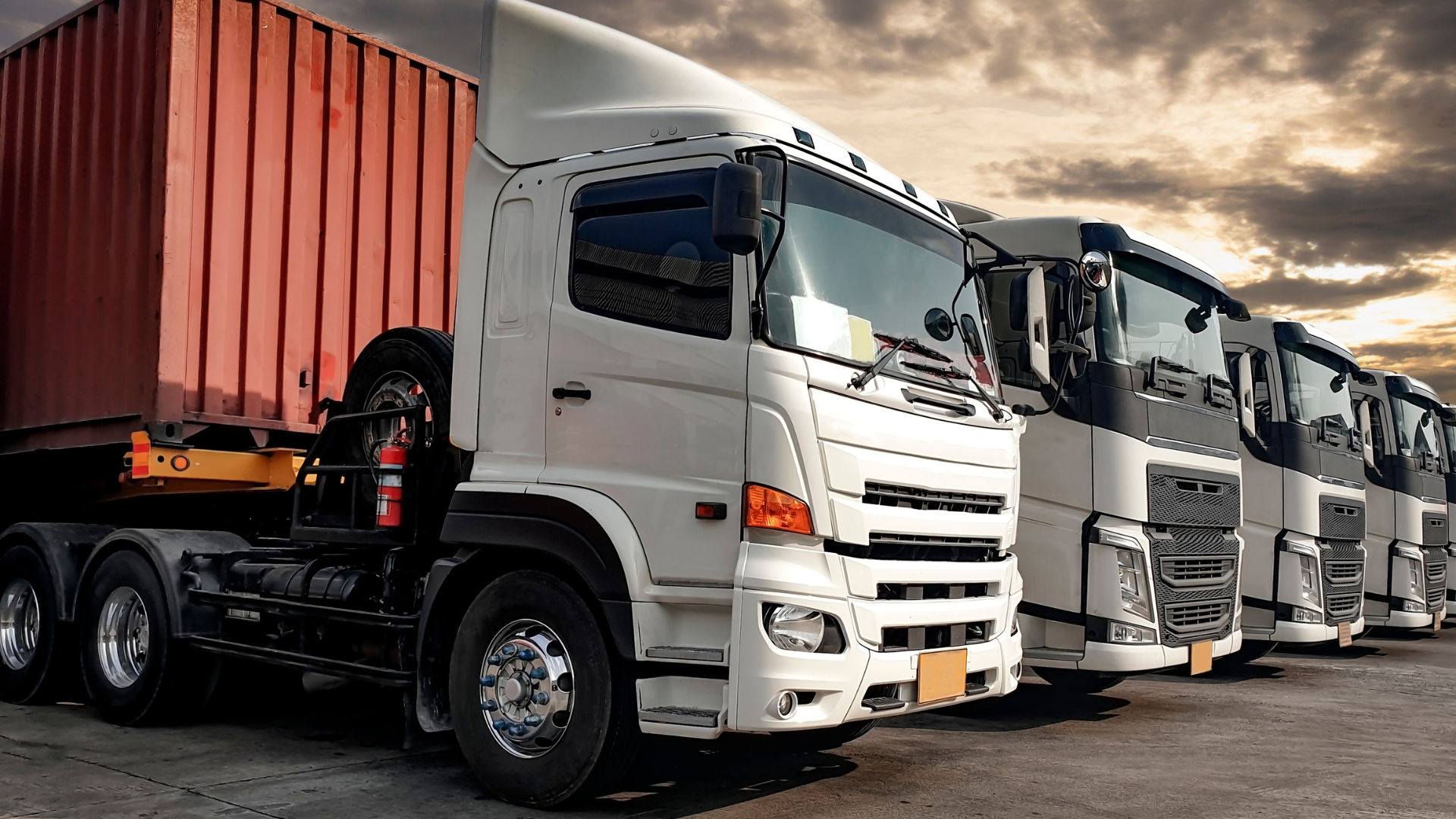 Trucks lined up in a row at a freight depot; white and red containers, cloudy sky.