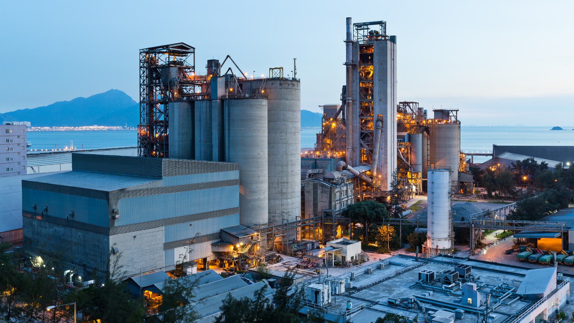 Cement factory at dusk with silos, towers, buildings, and coastal setting.