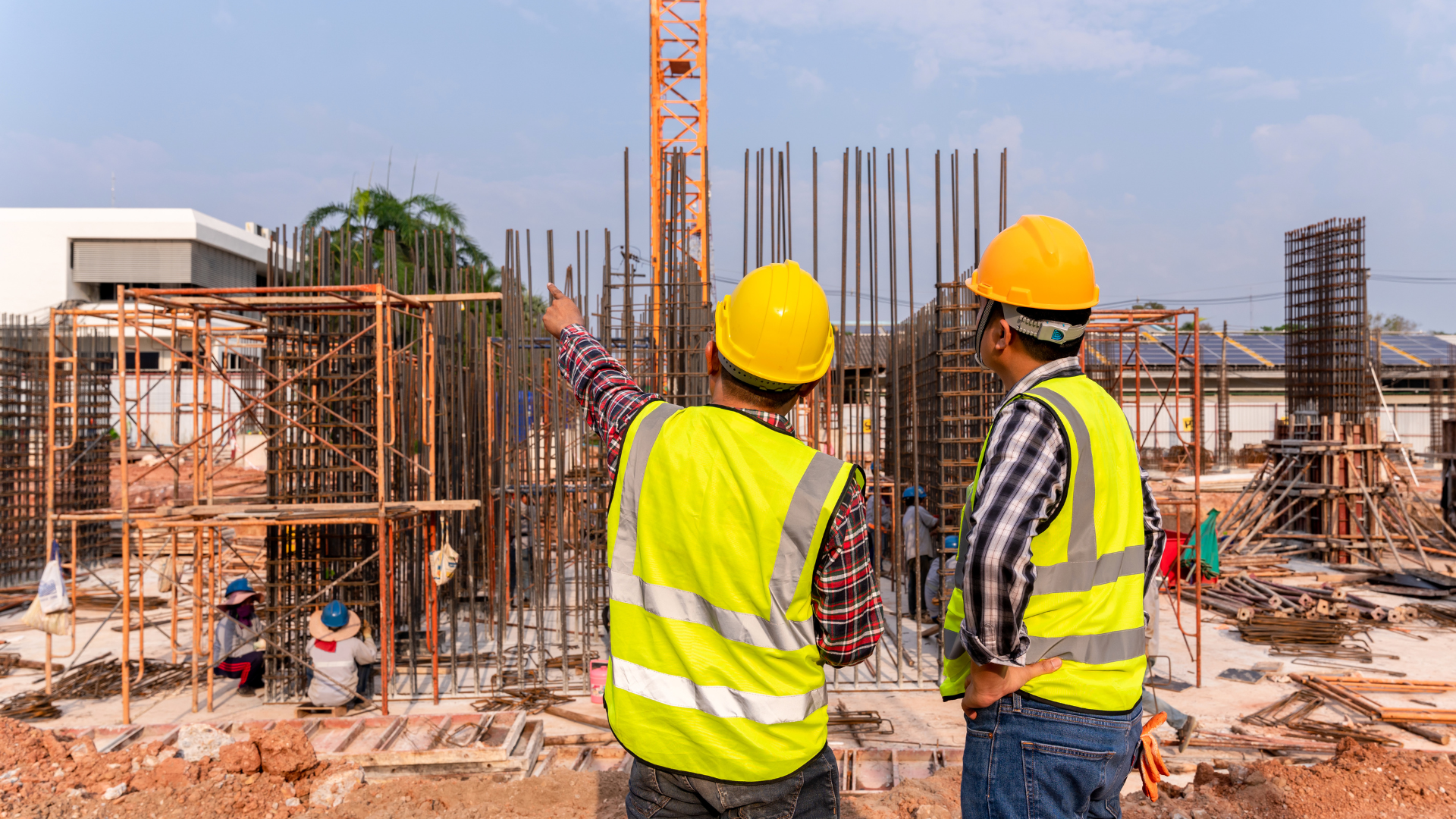 Two construction workers in yellow vests and hard hats inspect a building site, pointing at the structure.
