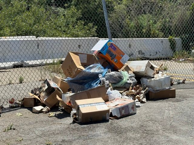 Pile of trash, including cardboard boxes and debris, on pavement by a chain-link fence.