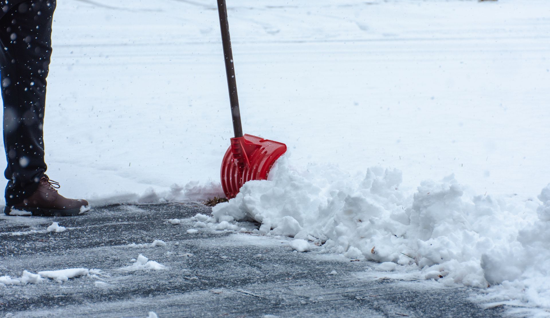 A person is shoveling snow from a driveway with a red shovel.