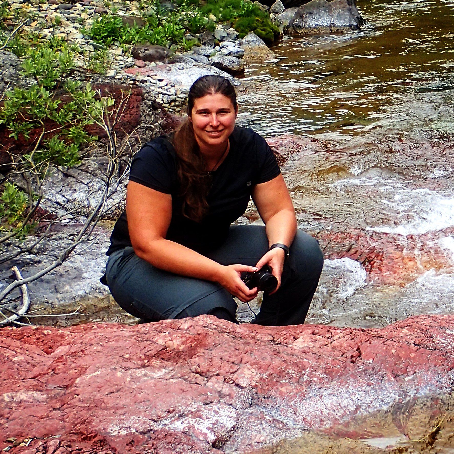 A woman is kneeling on a rock near a river holding a camera