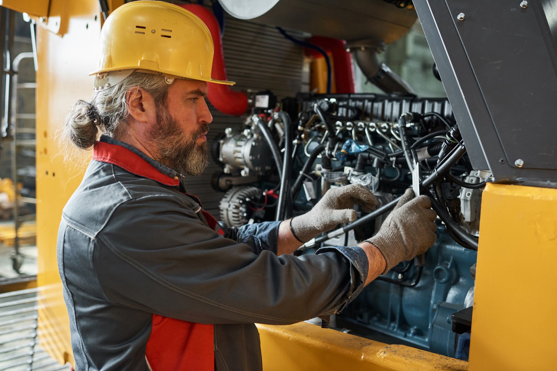 Mechanic in yellow hard hat repairs machinery engine, using tools. Inside a workshop.