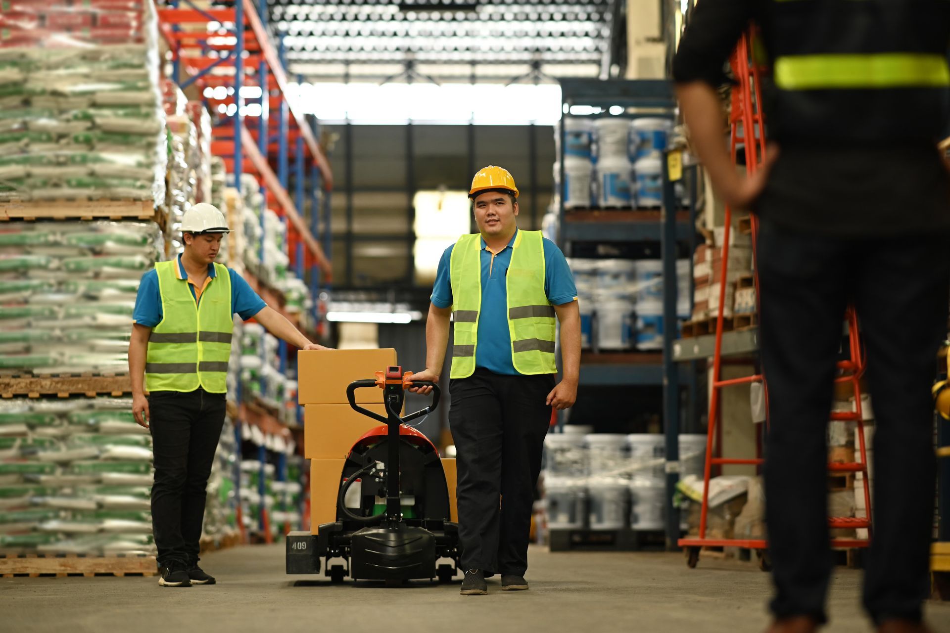 Warehouse workers with pallet jack and safety vests in aisle.