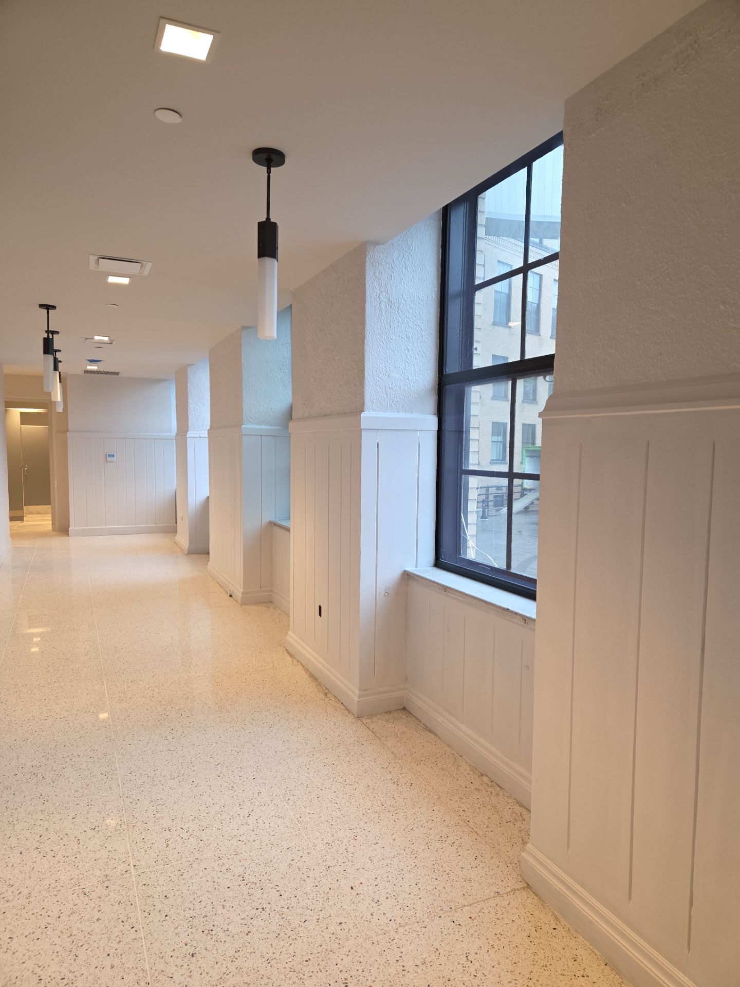 Hallway with white walls, terrazzo floor, window, and pendant lights.
