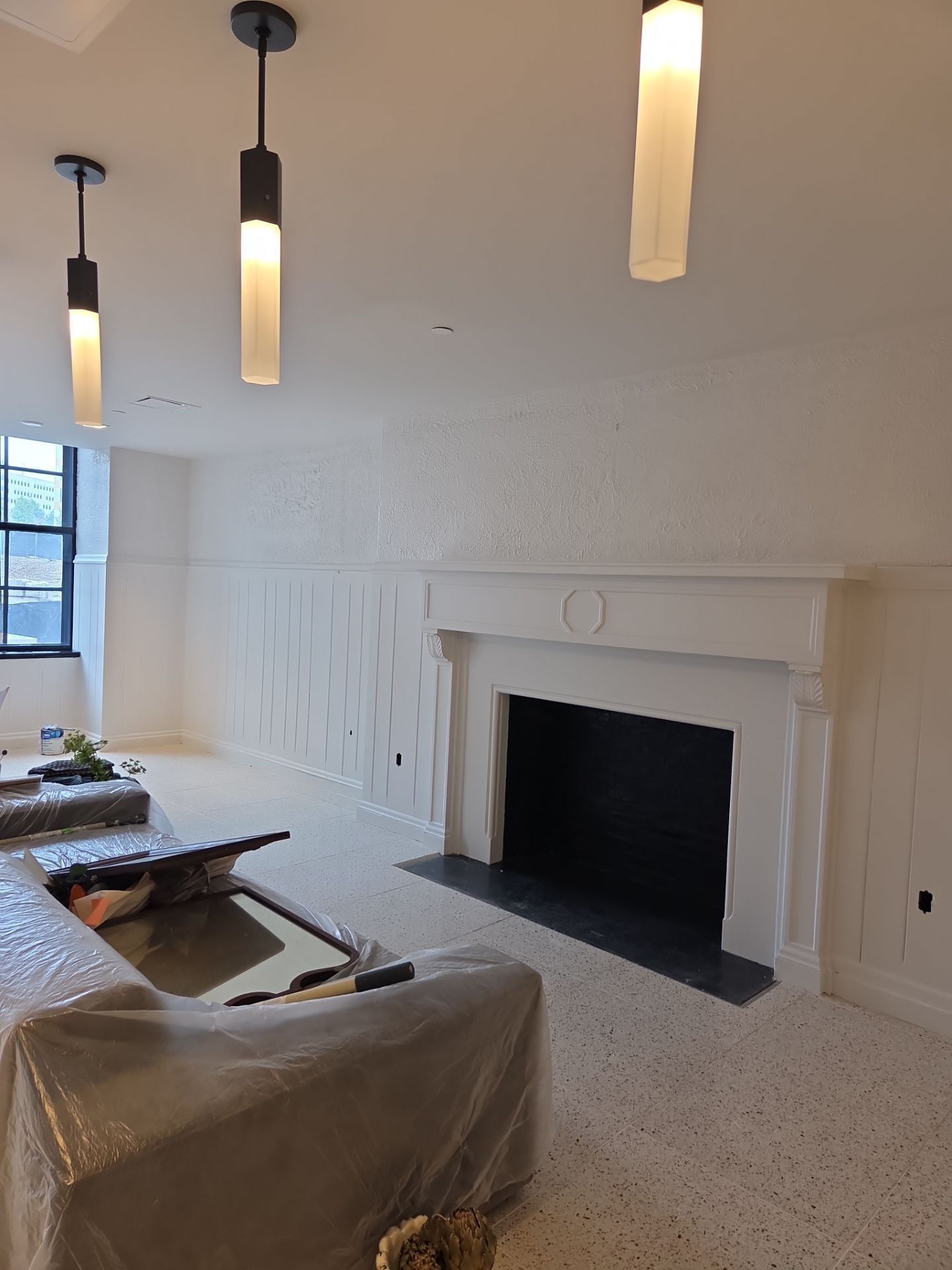 Living room with fireplace, white walls, pendant lights, and speckled flooring. Covered furniture in foreground.