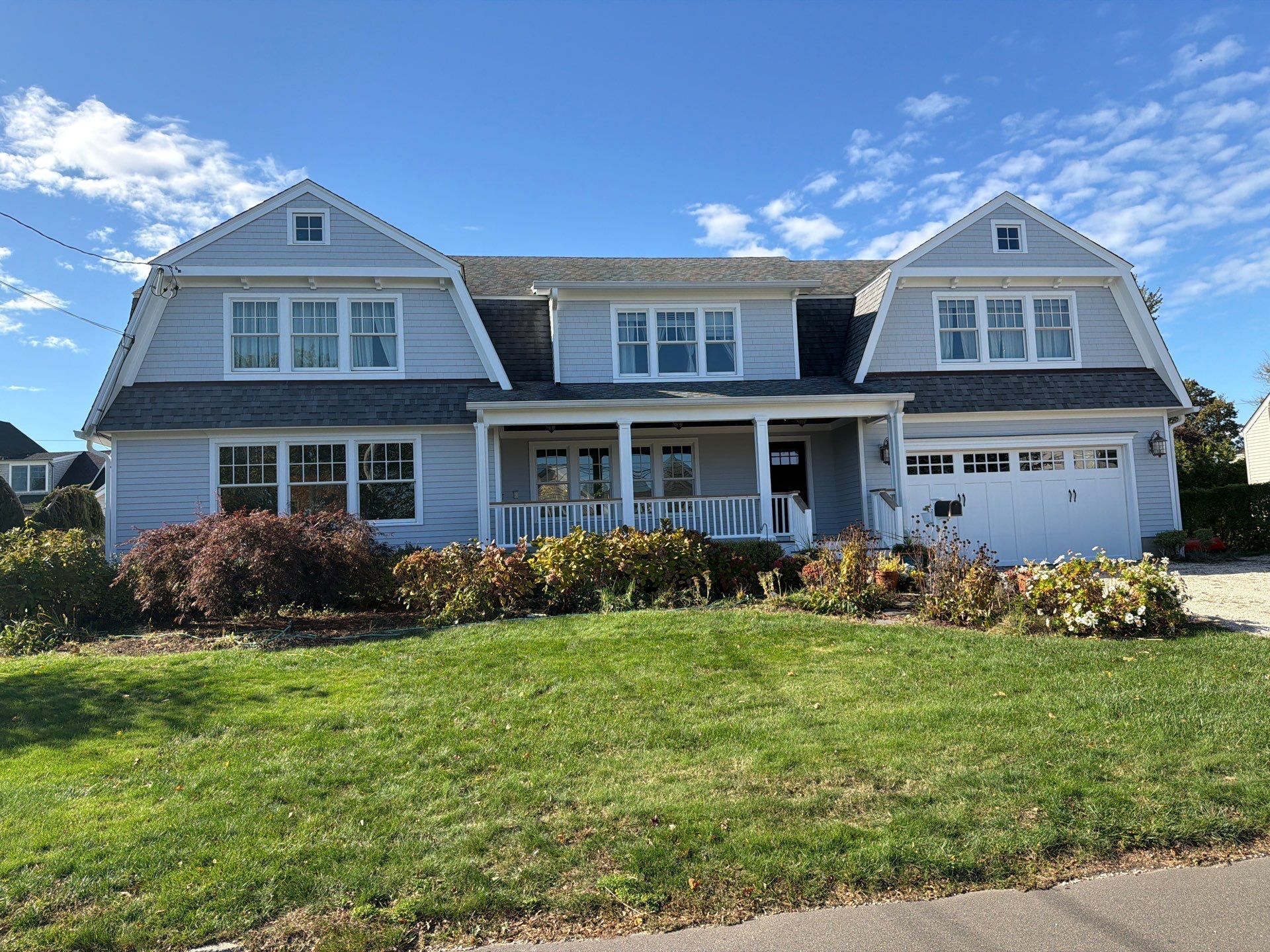Two-story gray house with Dutch gambrel roof, white porch, attached garage, and green lawn under a blue sky.