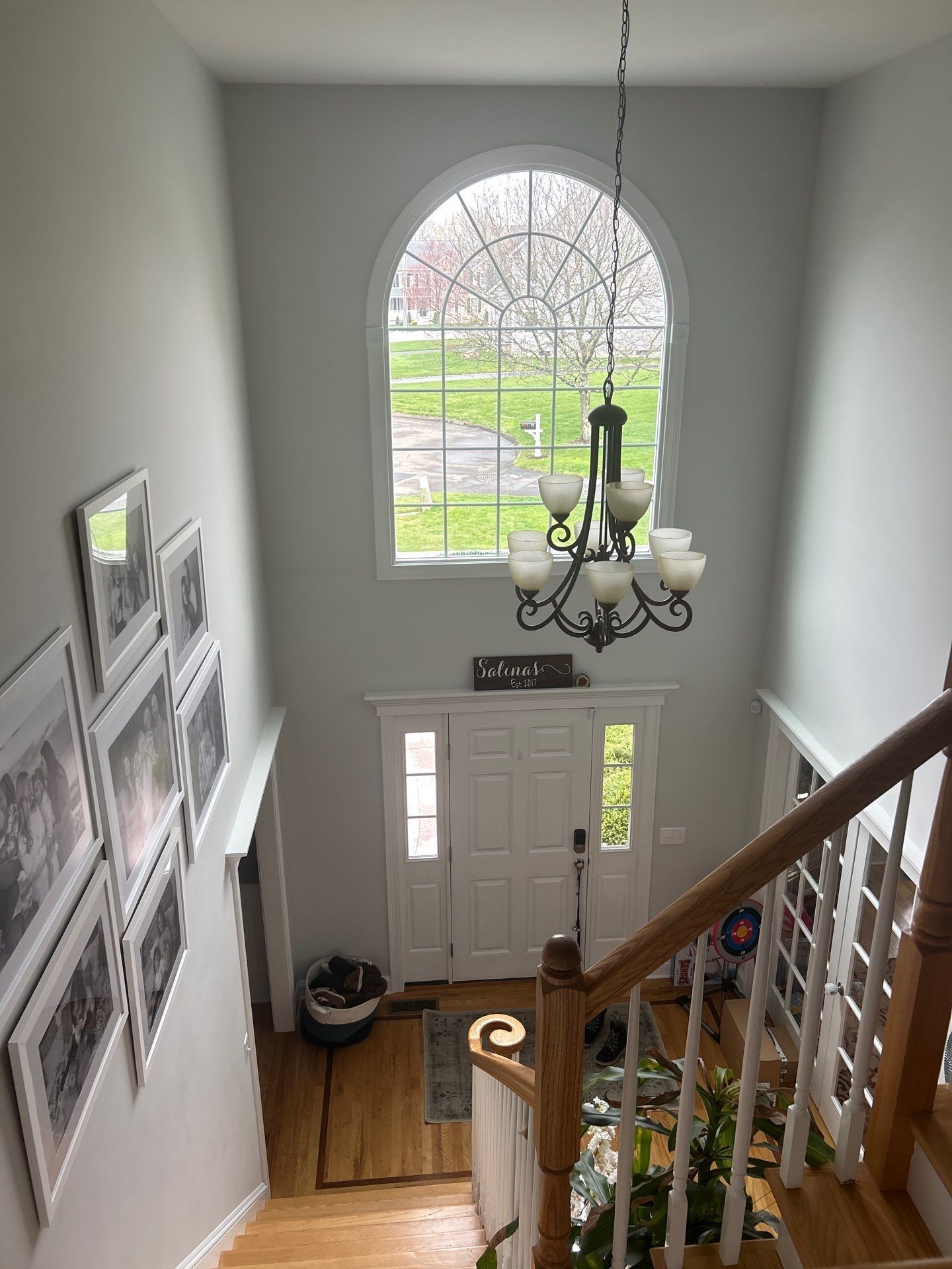 View from the top of wooden stairs, looking at a white front door and arched window. Gray walls and a chandelier.