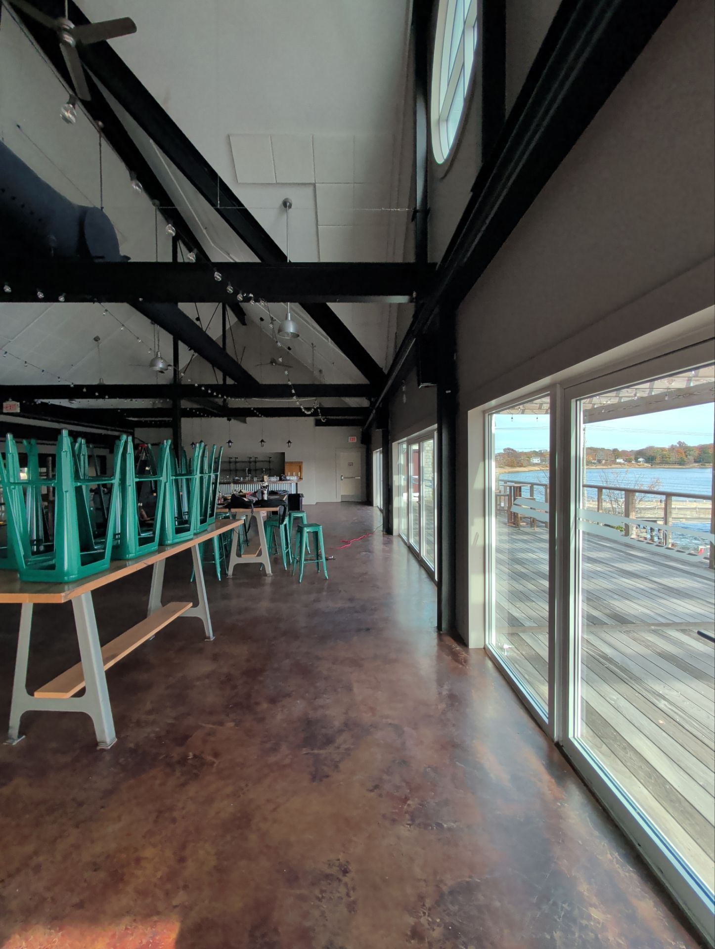 Interior of a building with tables, upside-down teal chairs, and large windows overlooking a dock. Brown floor.