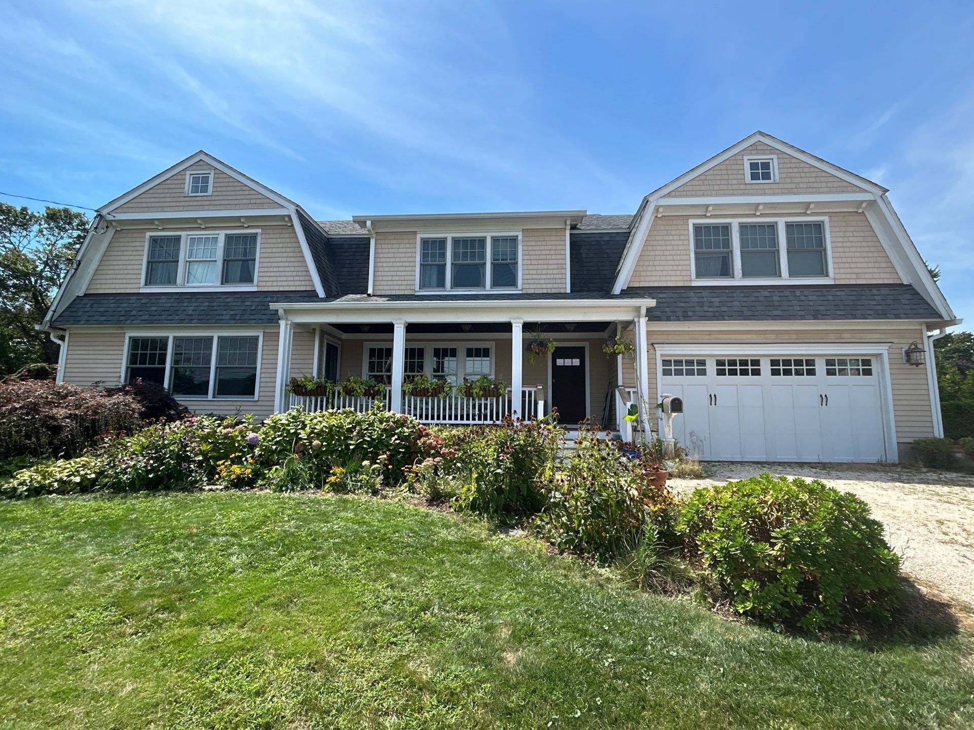 Tan house with dark roof, porch, garage, and front yard with flowers.