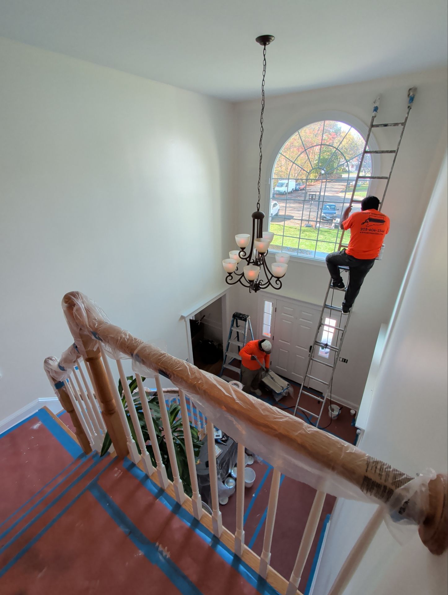 Two workers painting an interior stairwell. One on a ladder, another on the floor. High ceilings with a large arched window.