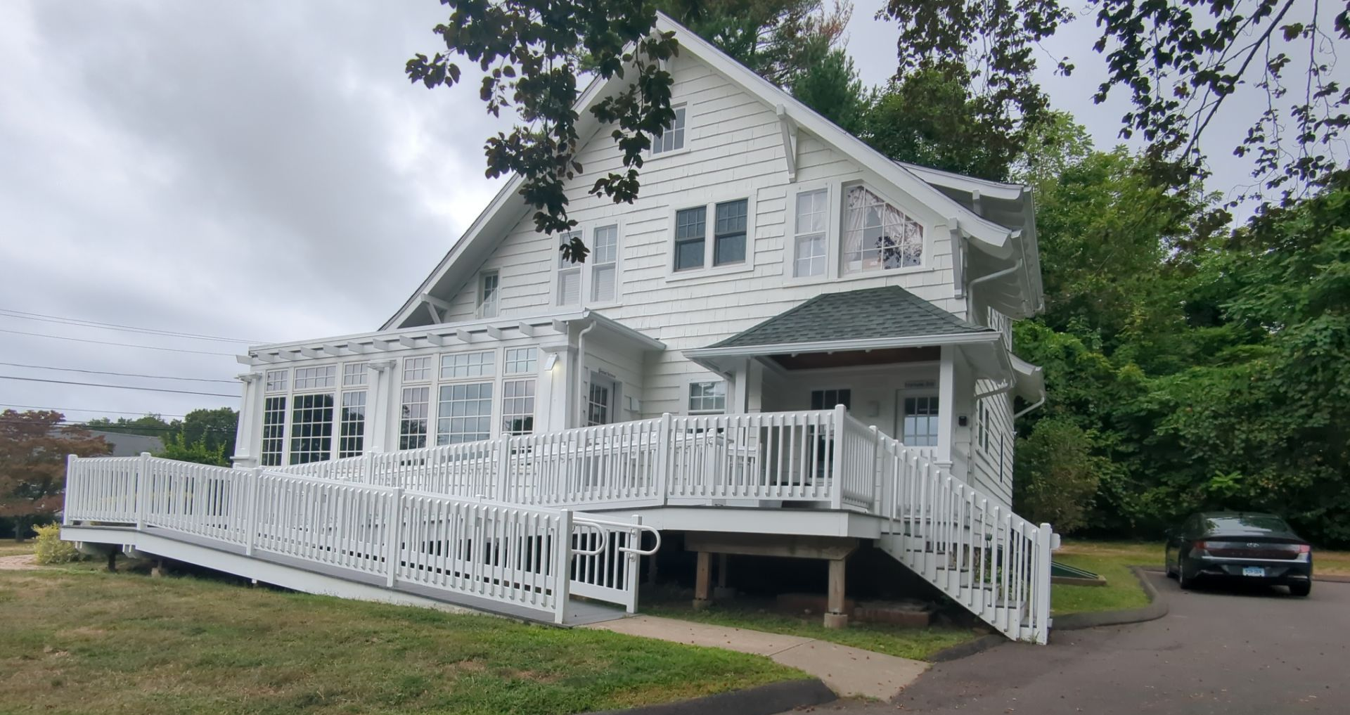 White house with wheelchair ramp, porch, and dark roof under cloudy sky.