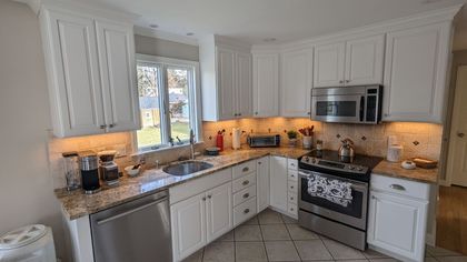 A bright, white kitchen with stainless steel appliances, granite countertops, and under-cabinet lighting. The sink is by a window.