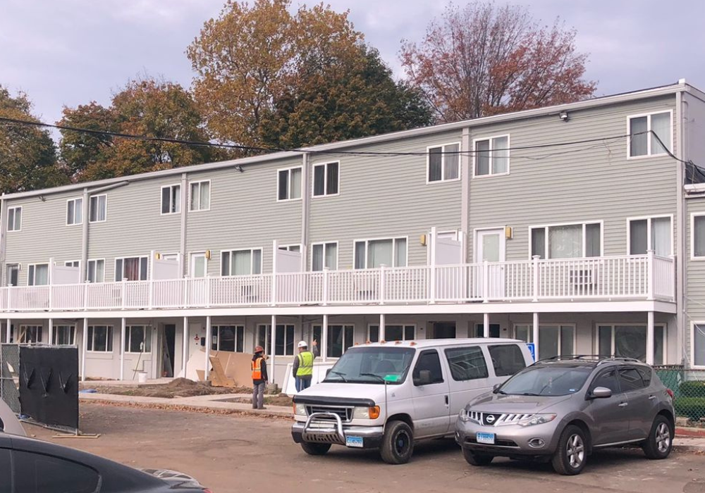 Apartment building with white balconies, gray siding, and vehicles parked in front under cloudy sky.