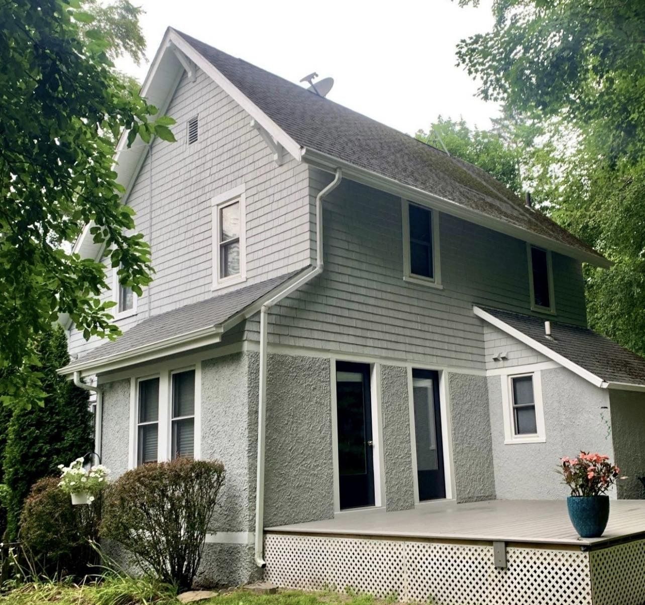 Two-story house with gray siding, white trim, and a porch, set amongst green trees.