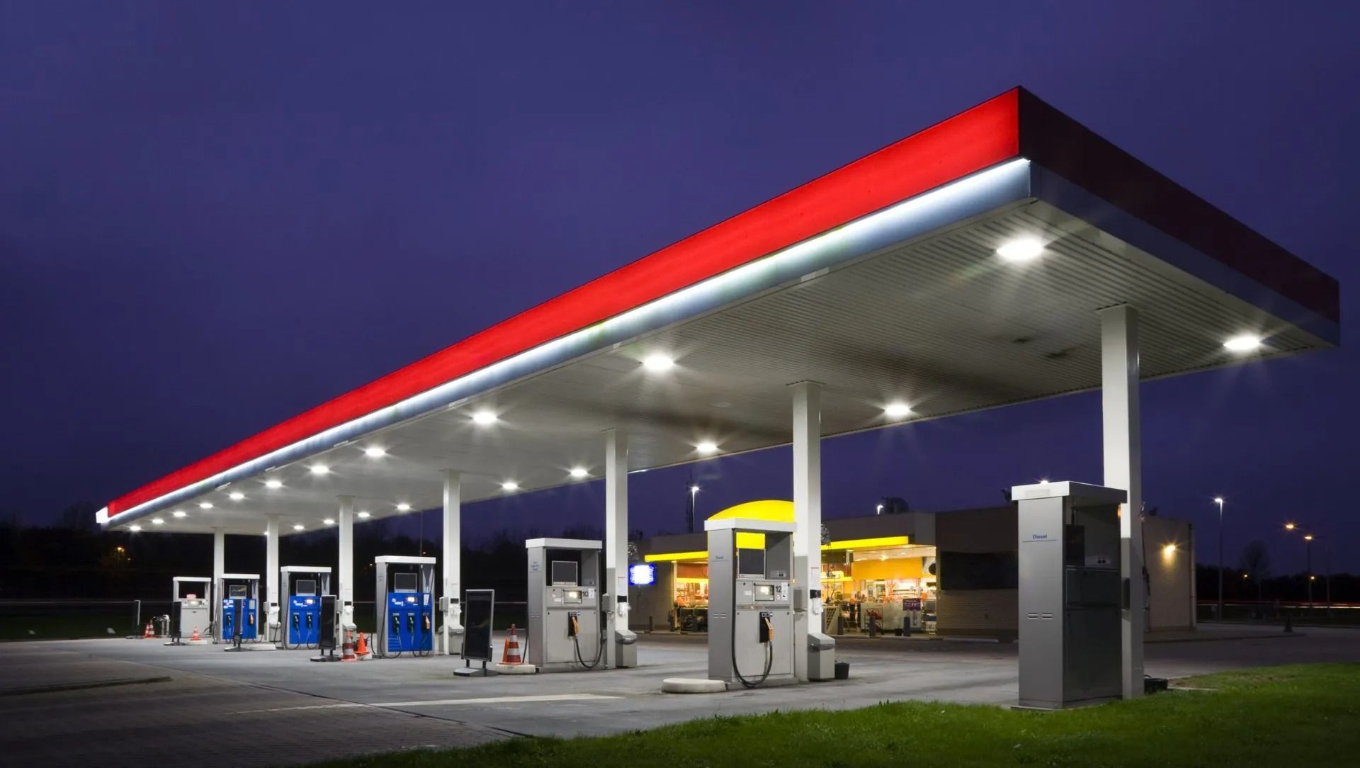 Gas station canopy illuminated at dusk, featuring red and white roof, fuel pumps, and store in background.