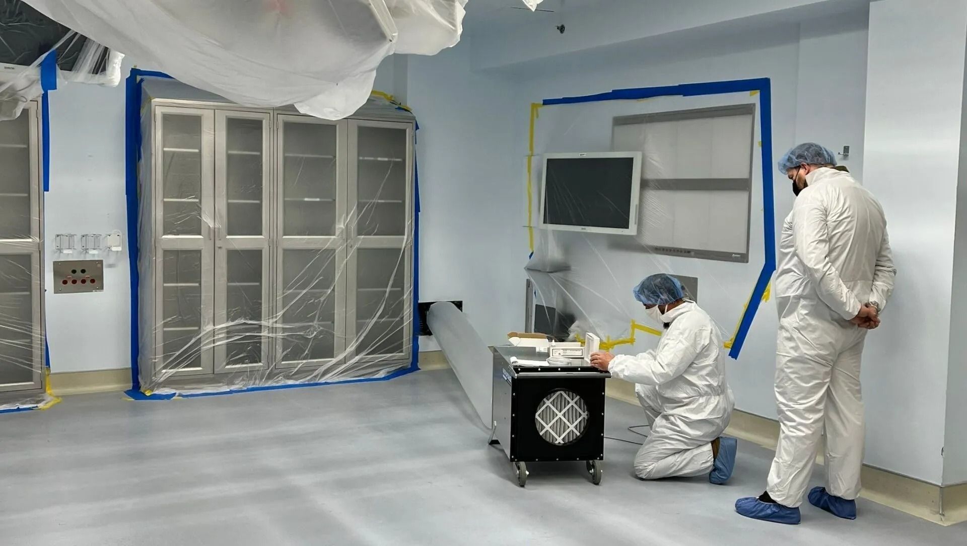 Two people in white lab coats work in a sterile room with stainless steel cabinets and equipment. The room is blue and white.