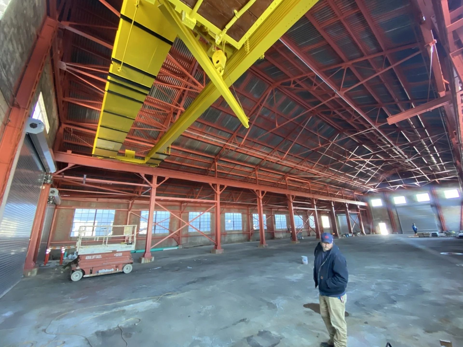 Inside a large warehouse with red support beams and a yellow overhead crane. A person stands in the foreground.