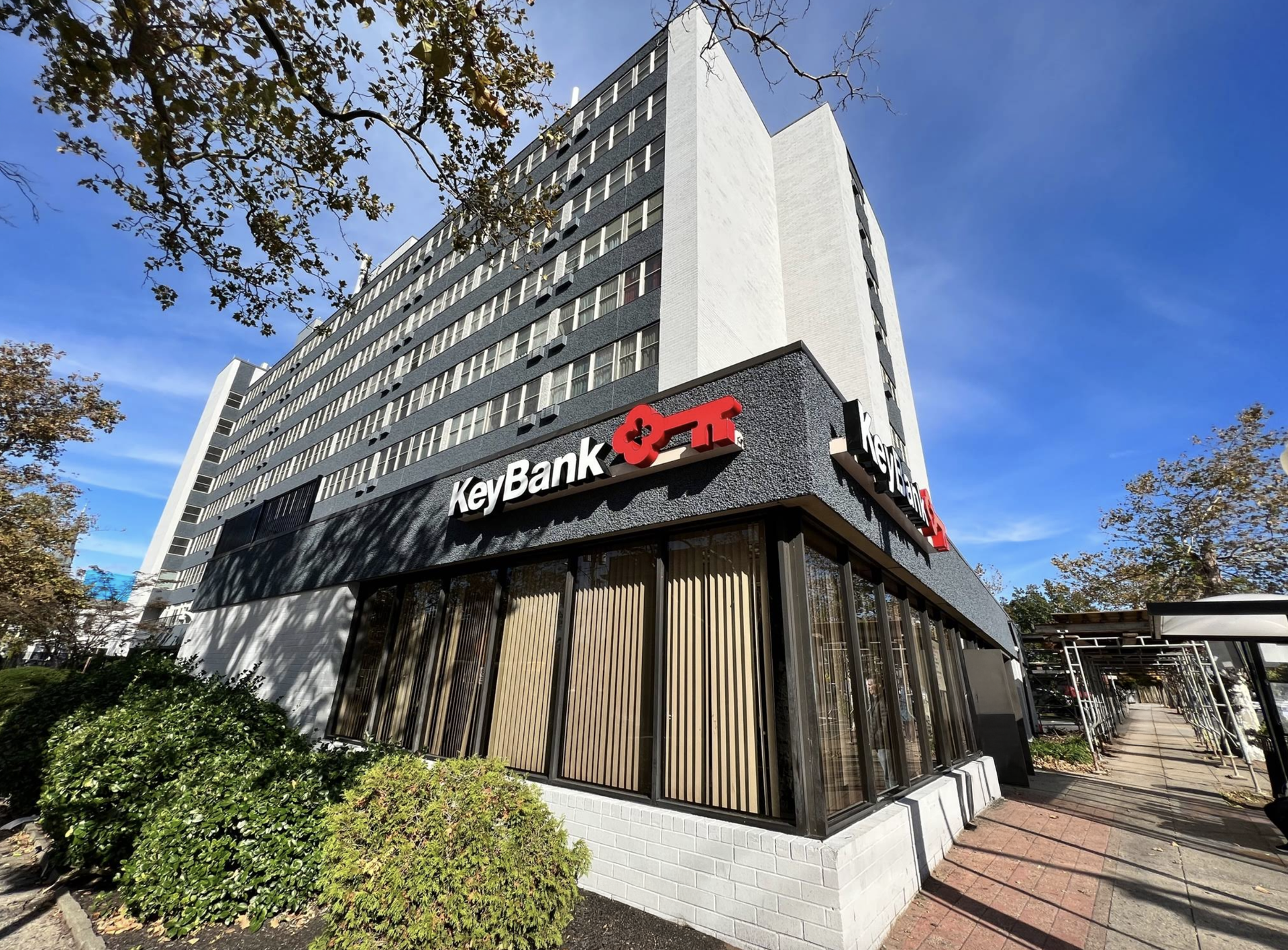 KeyBank building with red logo, tall, multi-story, glass windows, brick and shrubbery.