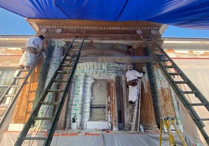 Two workers in protective suits on ladders are restoring a building facade. They are under a blue tarp, with visible damage and scaffolding.