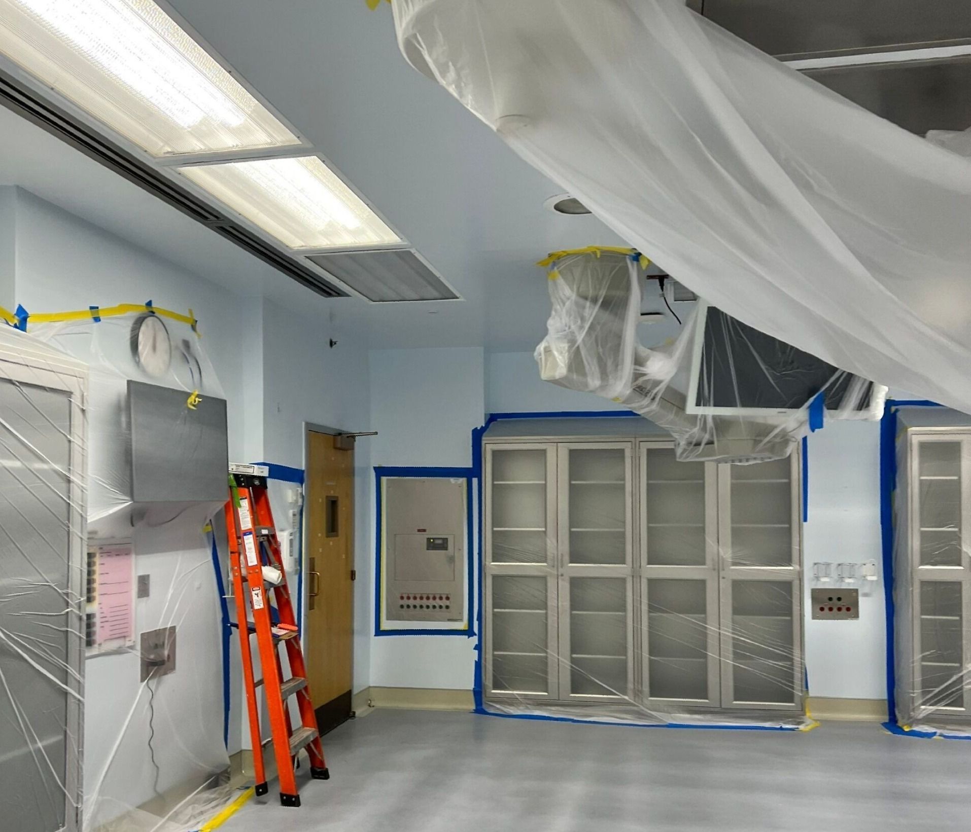 A medical room under renovation with protective plastic sheeting, masking tape, and a stepladder. The room has stainless steel cabinets, a light blue ceiling, and a gray floor.