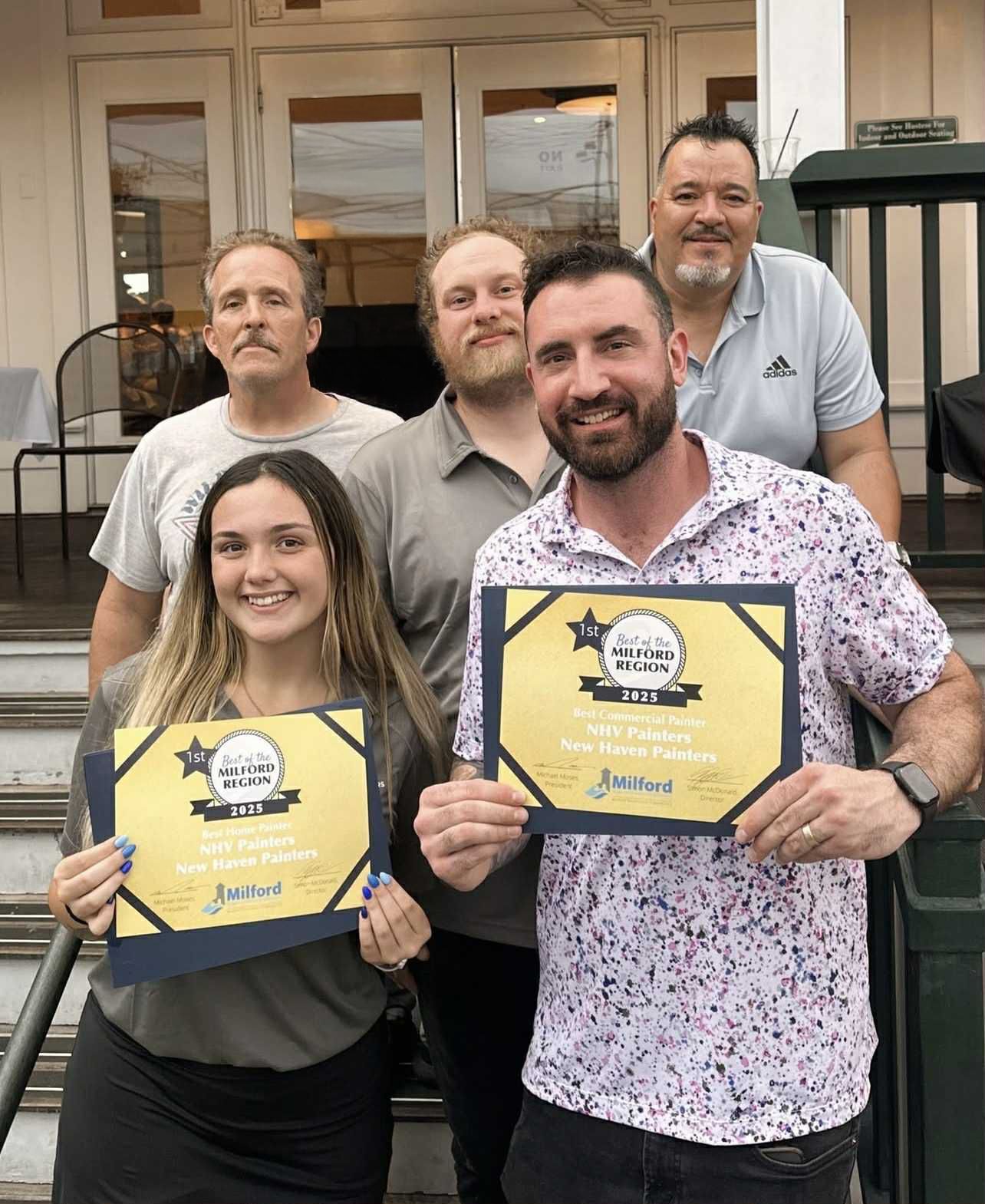 Group holding awards outside a restaurant. Two people in the front holding awards, smiles. Exterior shot.