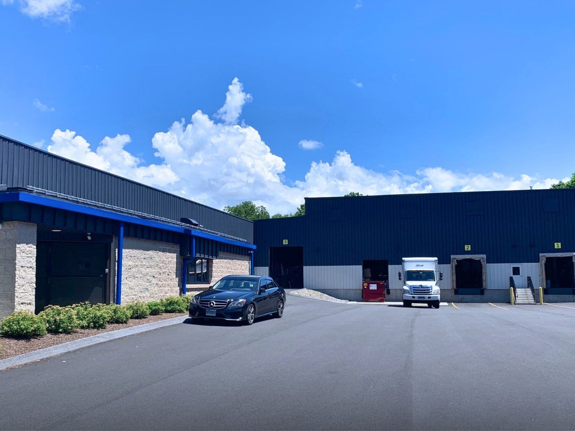 Exterior of a commercial building with loading docks, a car, and a truck on a bright, sunny day. Blue and gray tones dominate the facade.