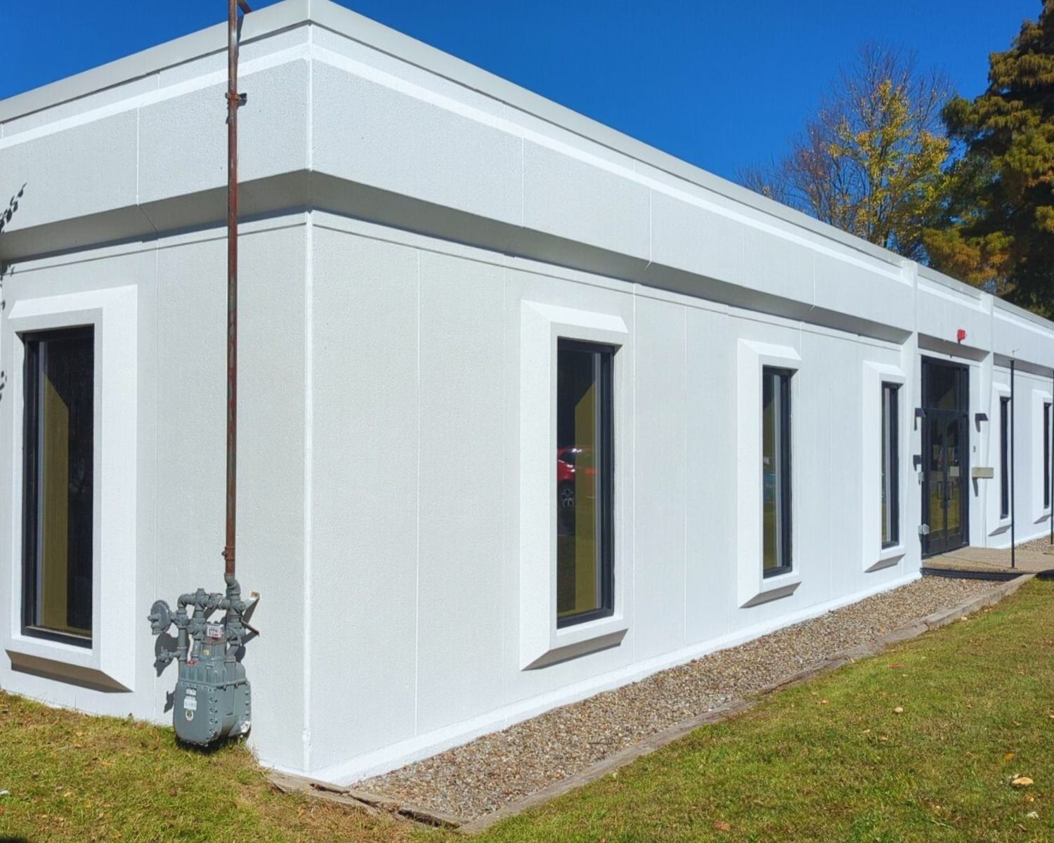 A one-story, light gray building with dark-framed windows and a gas meter, set against a grassy lawn and blue sky.