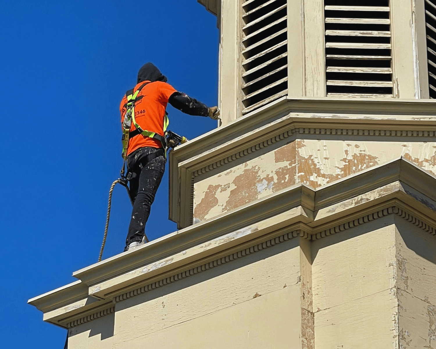 A person wearing a safety harness on a rooftop, painting the exterior of a light-colored building against a blue sky.