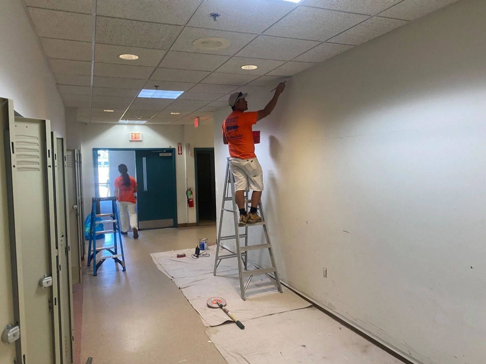 Two people painting a school hallway. One is on a ladder, painting the wall. The other walks further down the hall.