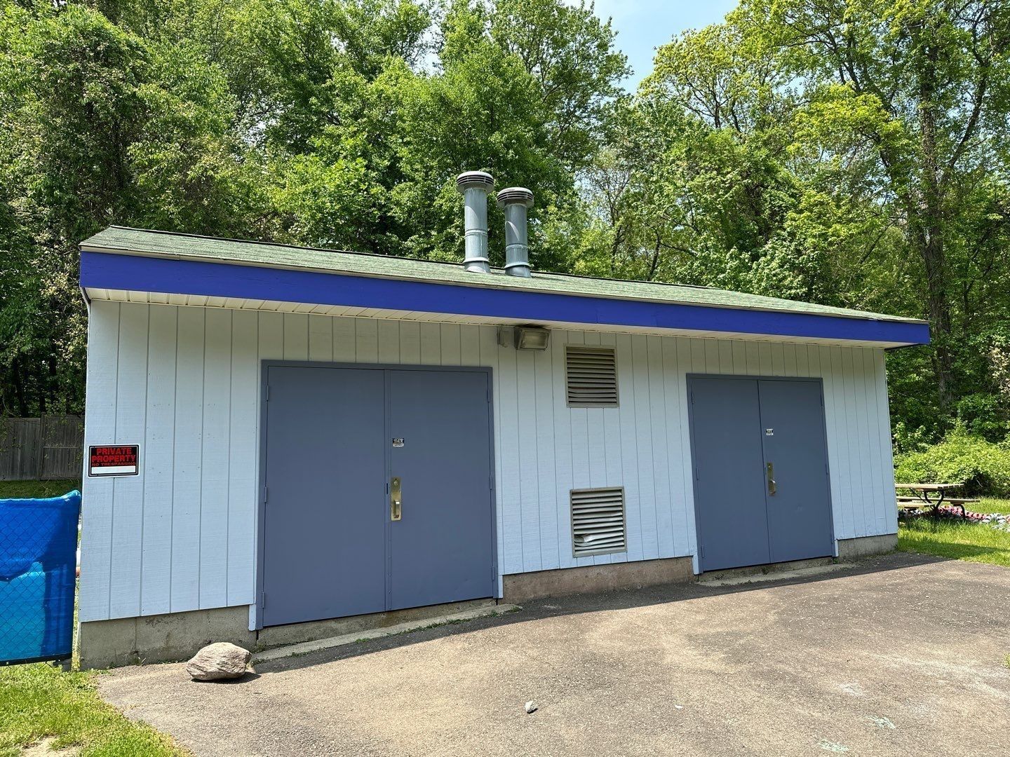 A white building with blue doors, a green roof, and two smokestacks stands against a backdrop of green trees.
