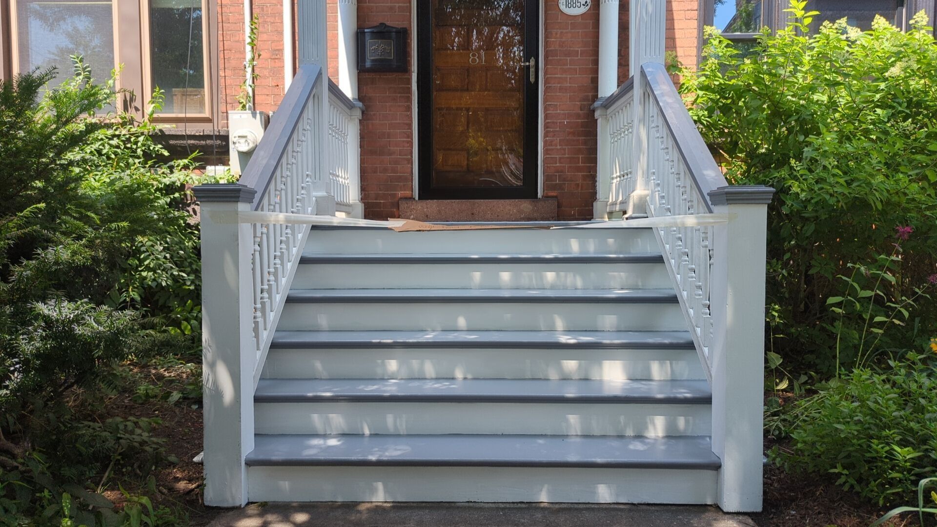 Grey and white painted stairs leading up to a red brick house with a black door, surrounded by green bushes.