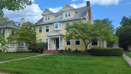 Yellow two-story house with a brick porch, green lawn, and blue sky.  A smaller house is partially visible on the left.