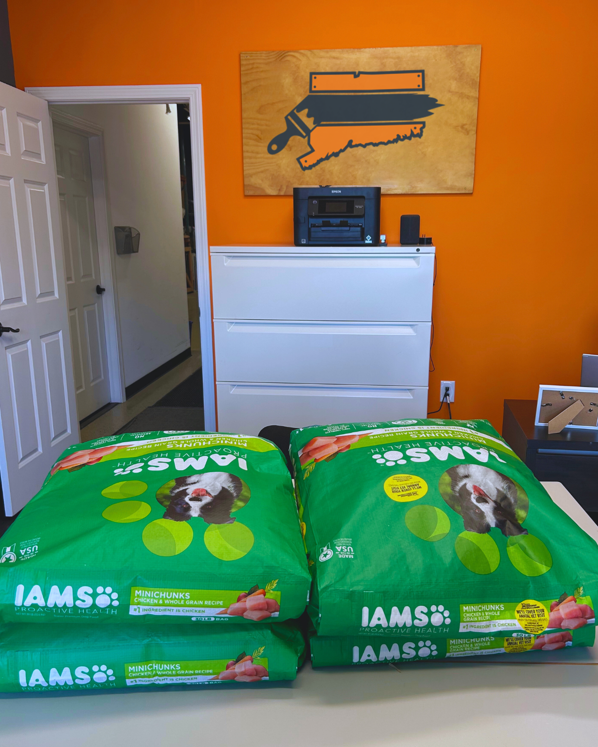Four large bags of IAMS dog food on a table, with a dresser and orange wall in the background.