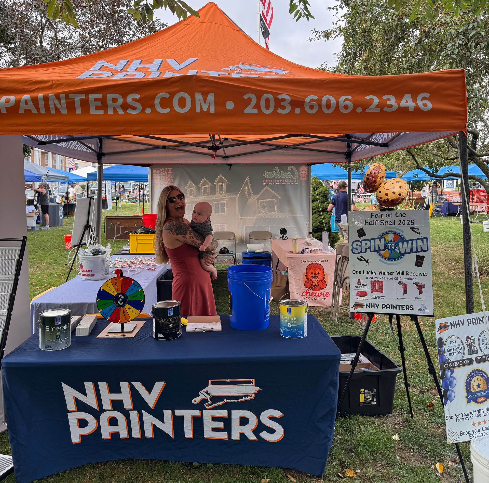 A woman holds a baby at a paint company booth with an orange tent.