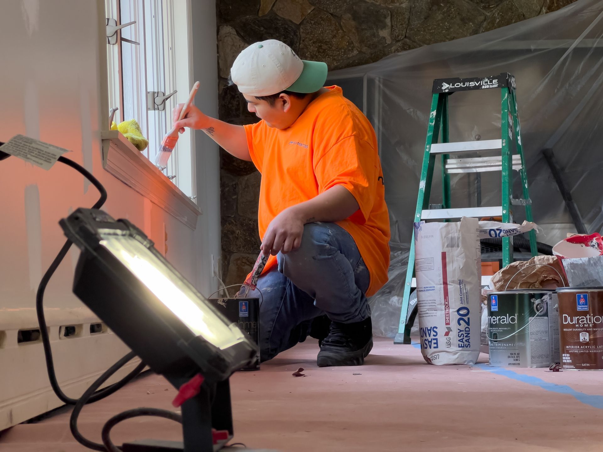 A man in an orange shirt is kneeling down while painting a window.