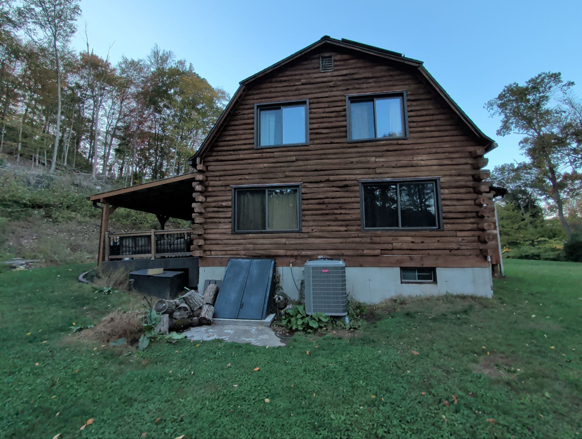 Log cabin with dark wood exterior, two floors, two windows on each level, and a porch. Green grass and trees surround it.