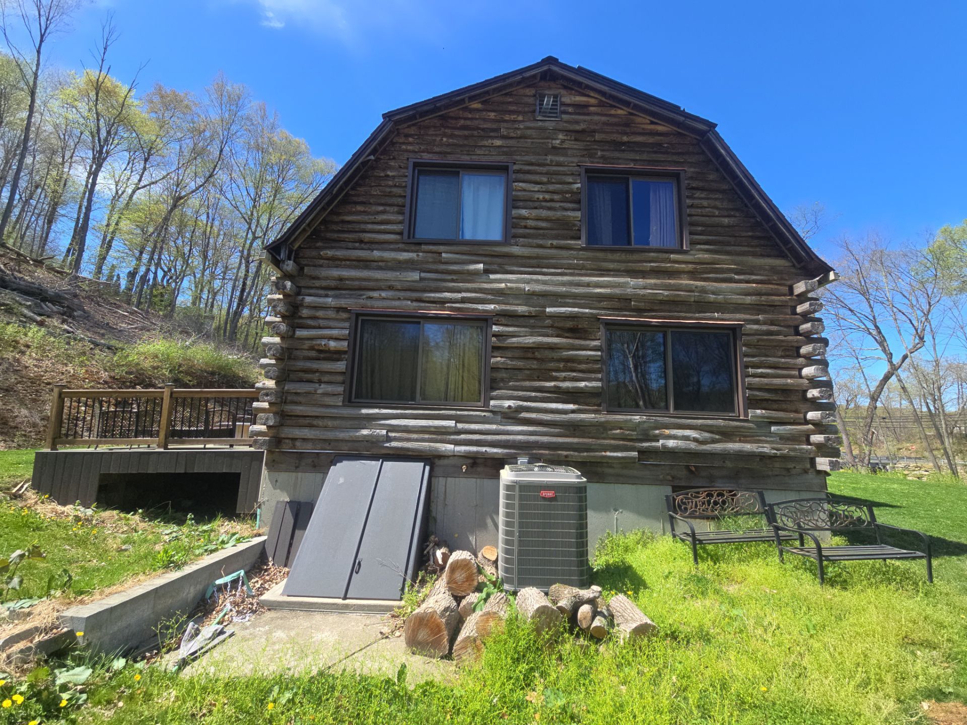 Log cabin with a deck on a grassy hill, a/c unit, and firewood.