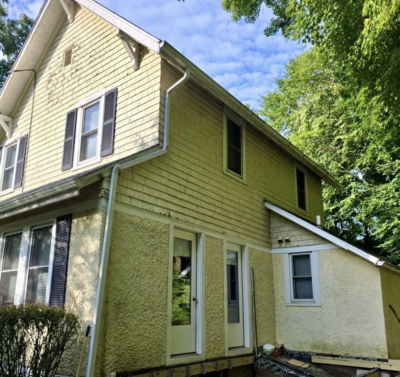 Two-story house with yellow siding, black shutters, and a small addition. Green trees surround the house.