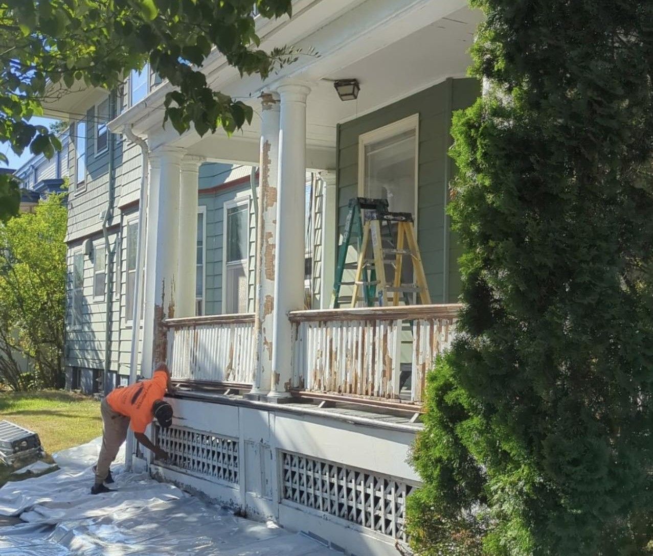 Person in orange vest works on a porch with peeling paint. White, green, and beige colors are visible.