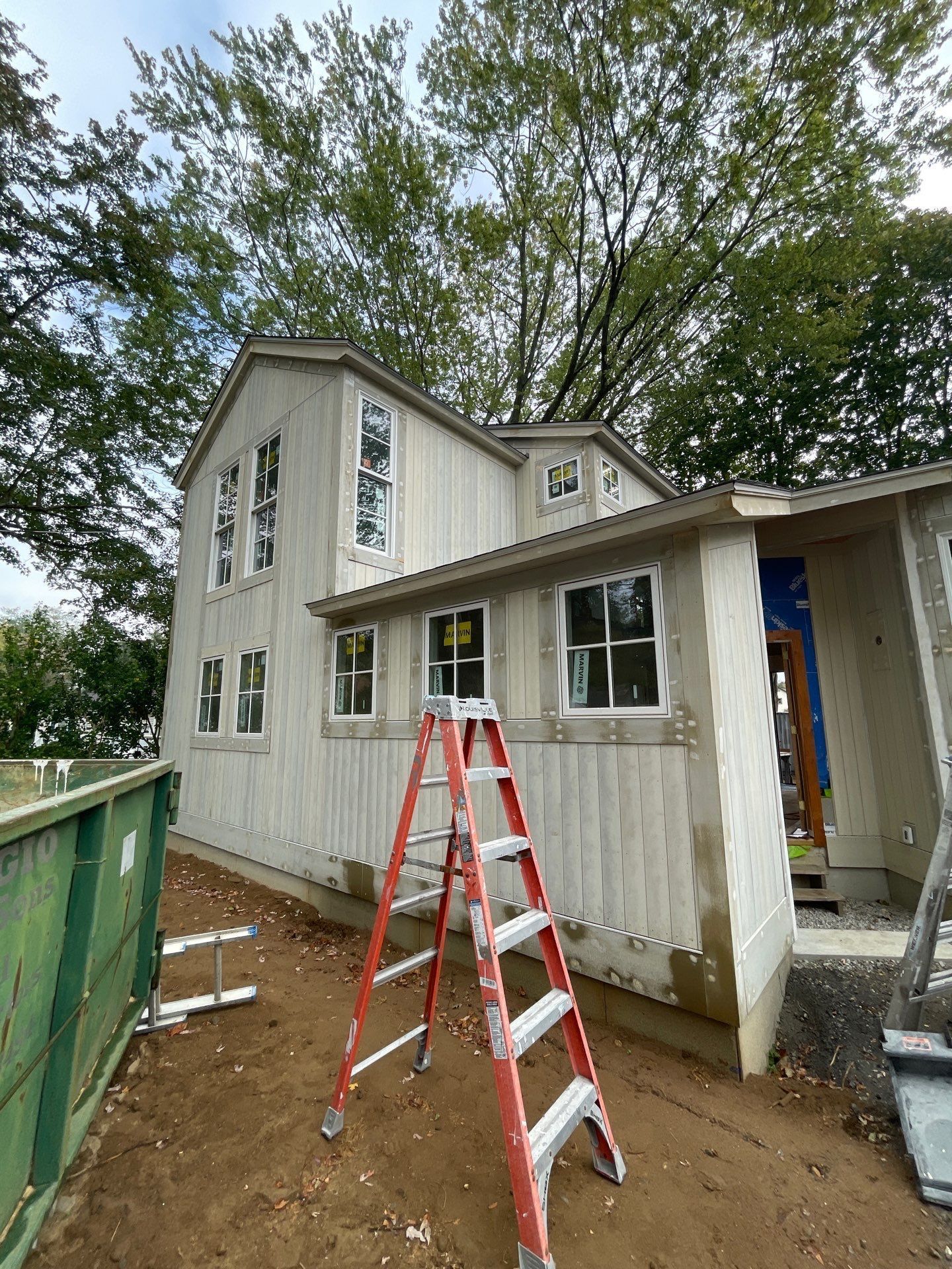 Construction site with a two-story building. A ladder stands against the building. A green dumpster is visible.