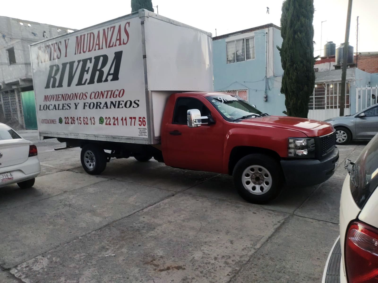 Camión de mudanzas rojo estacionado en una calle pavimentada con una caja de carga blanca; texto: