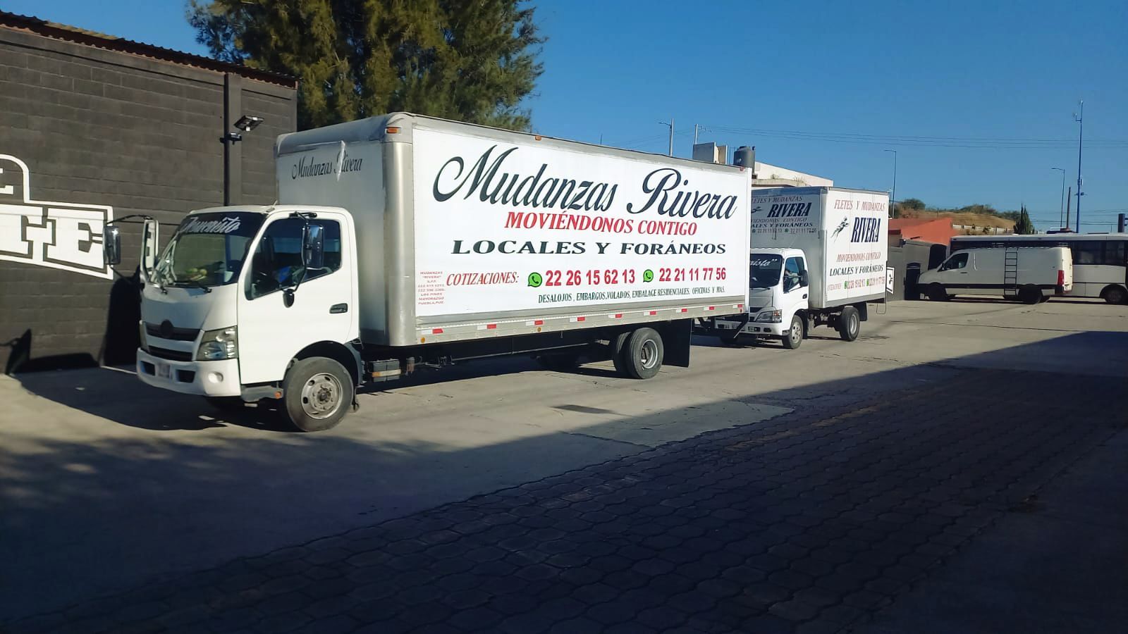 Camiones de mudanza blancos estacionados frente a un edificio en un día soleado. Los camiones tienen el nombre de la empresa en los laterales.