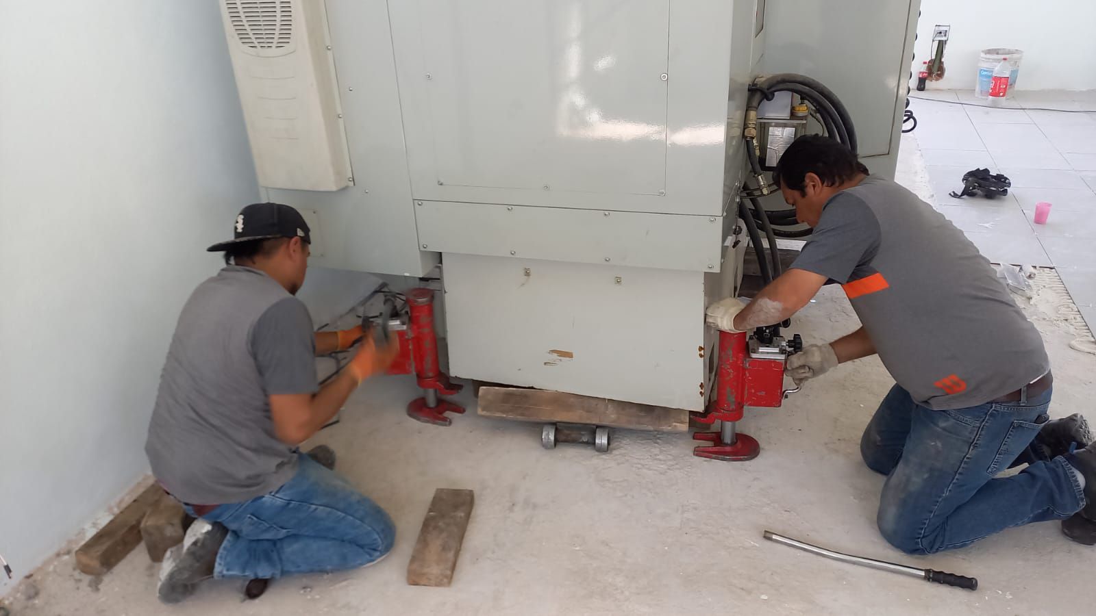 Dos trabajadores utilizan gatos para levantar un gran transformador eléctrico en un edificio.