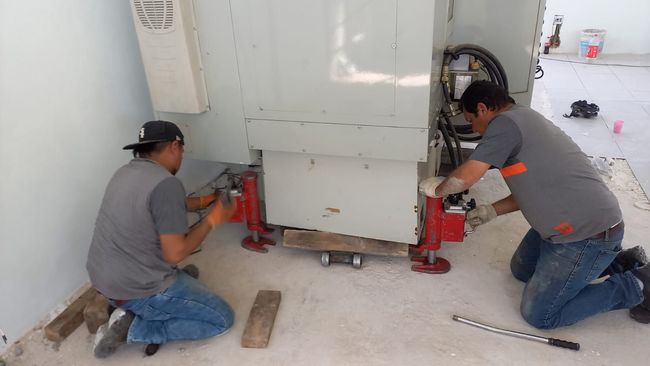Dos trabajadores utilizan gatos para mover una unidad eléctrica grande al aire libre.