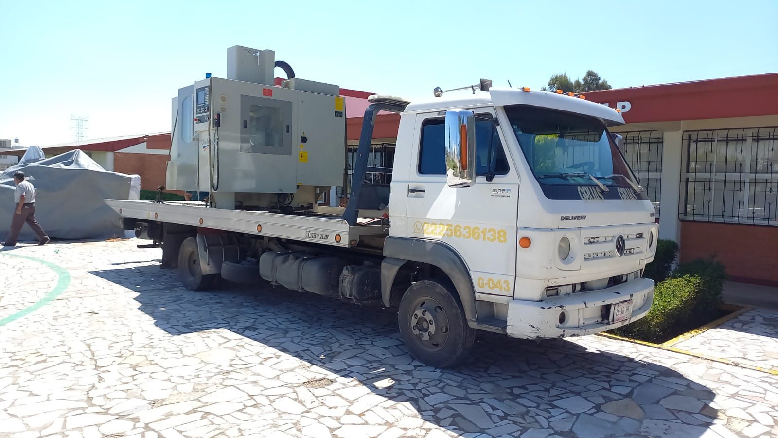 Un camión de plataforma con una máquina grande está estacionado frente a un edificio. Camión blanco, máquina gris.